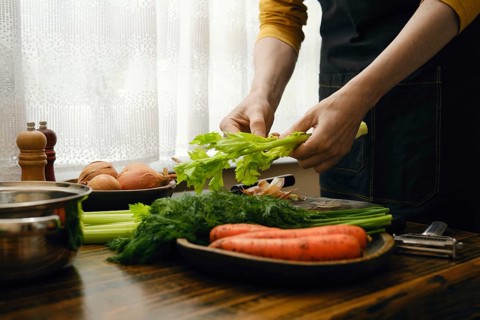 Person tears off a celery stalk. Other vegetables like carrots and onions are nearby. Light comes