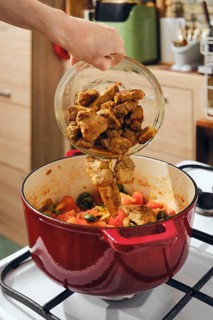Person is pouring fried pork pieces into pot filled with fresh vegetables on stove. Process of