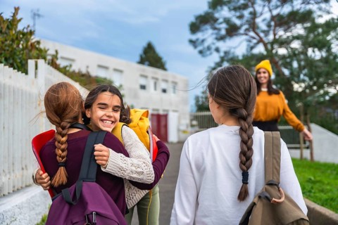 Two cheerful schoolgirls with backpacks hug joyfully outside a school building as a smiling adult
