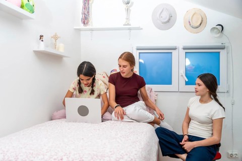 Teenage sisters or friends relaxing in a cozy bedroom, interacting and sharing a laptop while
