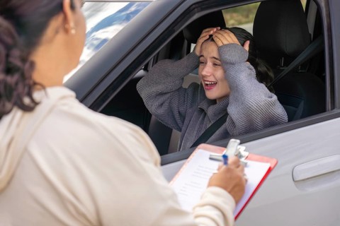 Young woman student holding her head and laughing while driving instructor writes evaluation notes,