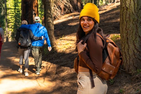 Woman in a yellow beanie and backpack smiling at the camera as friends walk ahead on a sunny trail