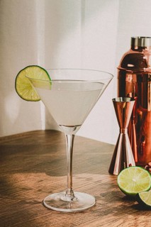 A clear cocktail sits in a glass with a lime slice on the rim. A copper shaker and halved lime are