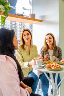 Three young women smiling and chatting over coffee and brunch at a sunny cafe window table,