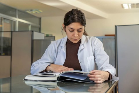 Young female scientist in white lab coat sitting at a glass table intently reading a large open
