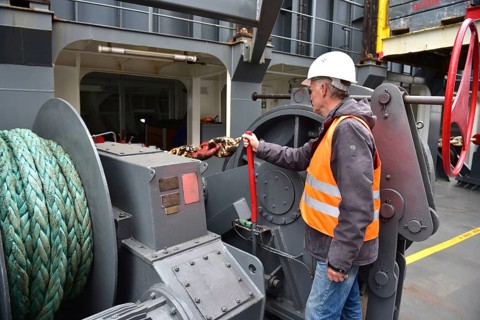 A worker with helmet and vest handles a rope on a large machine on a ship, worker with the anchor