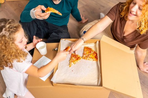Family members eating hot dogs pizza from a cardboard box, celebrating their relocation into a new