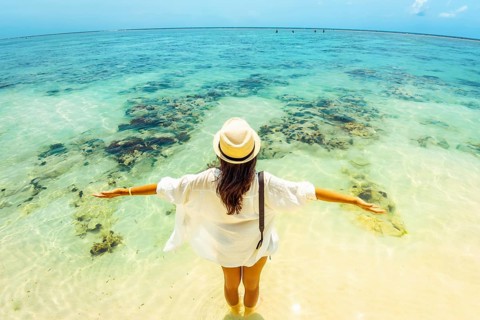 Young carefree woman wear straw hat stands arms wide open hug ocean in shallow, crystal-clear