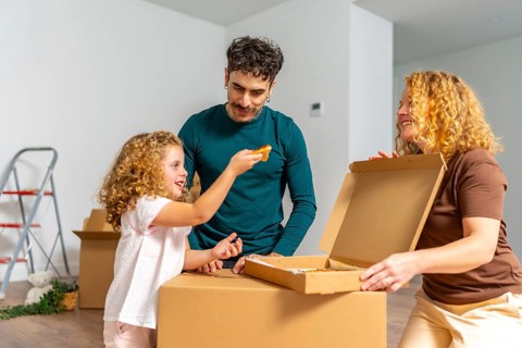 Happy family celebrating moving into their new home, sitting on cardboard boxes and sharing pizza