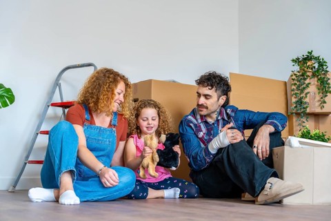 Happy family parents and daughter sitting on the floor amid moving boxes, smiling and celebrating