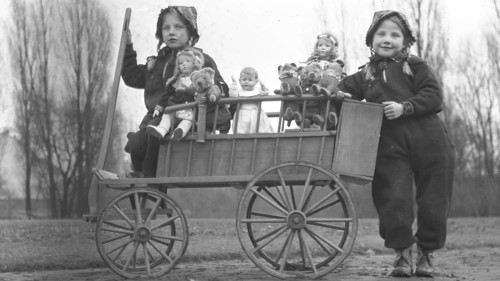 Historical black and white photo of two children with a wooden handcart, dolls and small stuffed