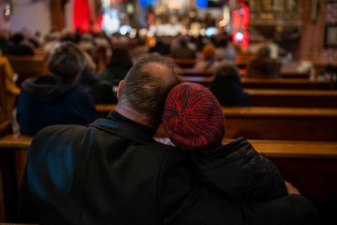 View from behind of a couple sitting closely in wooden church pews during a service, with the woman