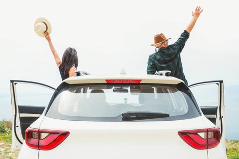 Couple cheers with arms raised while standing by white car overlooking coastal cliff. Bright