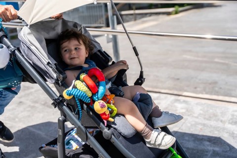Curious baby boy sitting in a stroller with a colorful toy, smiling and looking away during a sunny