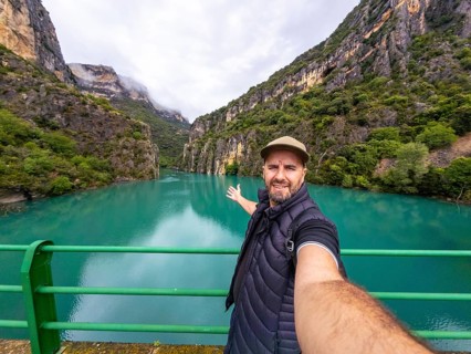 Man on a bridge taking a selfie and pointing across the turquoise cinca river flowing through the