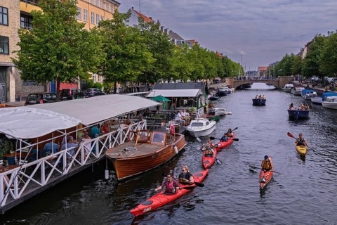 Menschen paddeln in Kajaks auf einem städtischen Kanal mit umliegenden Bäumen und Gebäuden,