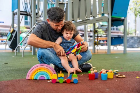 Father and son sit on soft playground flooring, playing with colorful wooden and plastic toys,