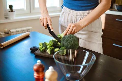 Woman prepares smashed broccoli with cheese in bright kitchen. Woman cuts fresh broccoli on cutting