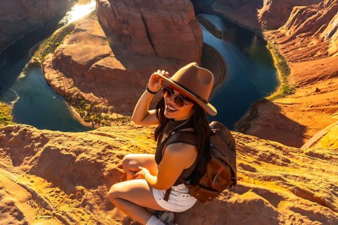 Woman in hat and sunglasses sits on sandstone cliff edge smiling at camera, overlooking the