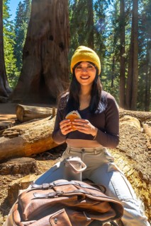 Young woman smiling while sitting on a large fallen sequoia log, eating a sandwich and a drink in a