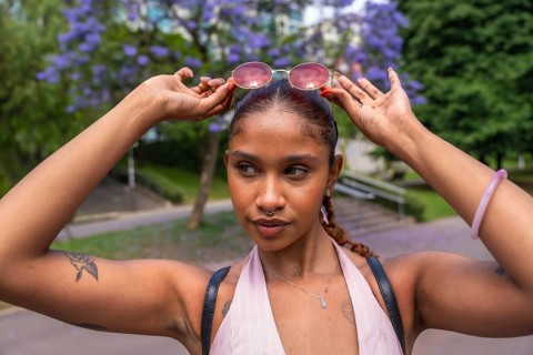 Natural young woman with dark skin and braided hair raising pink sunglasses on her head, posing