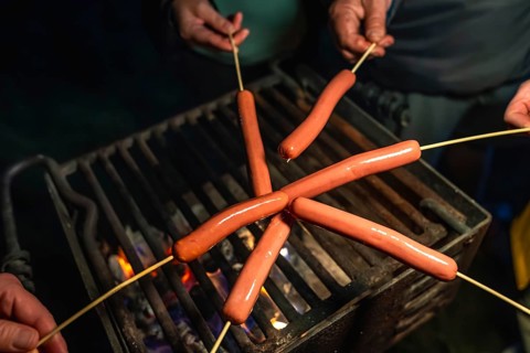 People holding skewers with hot dogs over a glowing campfire grill grate, cooking and enjoying a