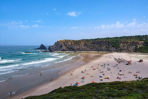A busy beach with colorful umbrellas and rock cliffs under a blue sky, Odeceixe, Faro, Portugal