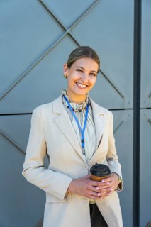 Young businesswoman smiling, holding a takeaway coffee cup, wearing a smart blazer and lanyard,