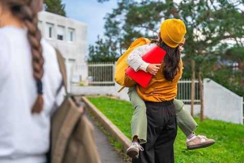 Mother carries her smiling daughter on her back after school, sharing a joyful piggyback hug as