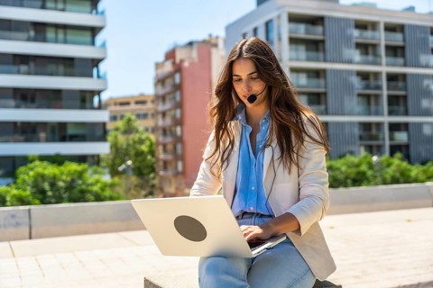 Woman wearing a headset, typing on a laptop, and conducting business calls while working remotely