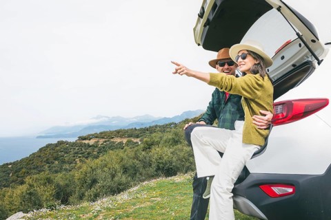 Happy couple sits in their white rented car trunk, laughing, enjoy nature views during spring road
