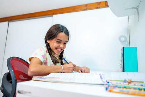 Happy schoolgirl with braided pigtails smiling as she studies and writes in a notebook at her white
