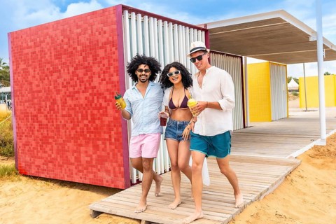 Diverse group of young friends is walking barefoot on a wooden boardwalk near colorful beach