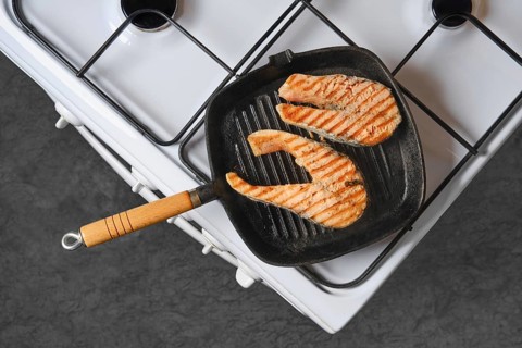 Salmon fillets are cooking on grill pan placed on stovetop. Grill marks are visible on fish.