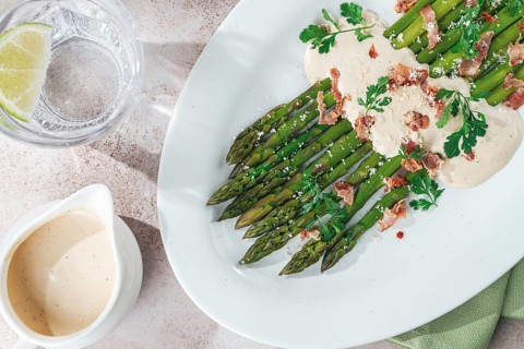 Fresh asparagus with cream sauce and bacon, on a white oval plate, light marble table, sunlight,