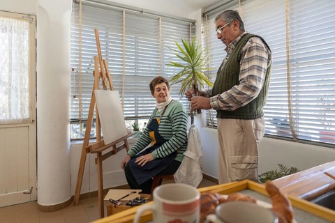 Wide shot of a senior woman seated at her easel laughing with her standing husband in a bright home