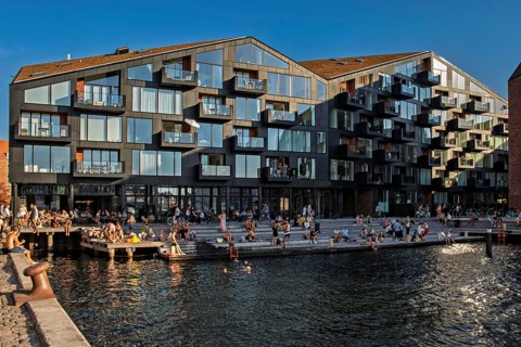 Menschen entspannen am Wasser vor einer modernen Glasfassade in der Abenddämmerung, Kopenhagen