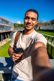 Young male student with dreadlocks and backpack, holding a reusable coffee cup and taking a selfie