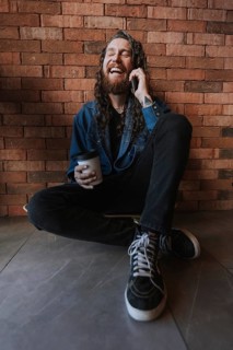 A man with long hair and a beard laughs heartily, sitting against a brick wall. He holds a phone to