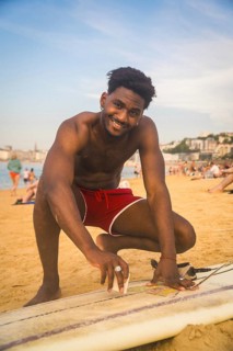 Young smiling man in red shorts applying wax to his surfboard on a sandy beach, preparing for a