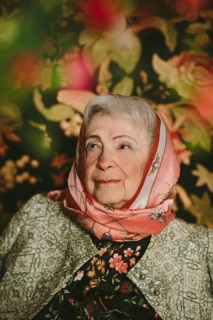 An elderly woman wearing a patterned silk scarf and jacket sits against a colorful floral backdrop.