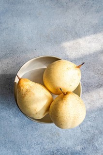 Three ripe Chinese white pears rest in a ceramic bowl placed on a textured light blue surface. The