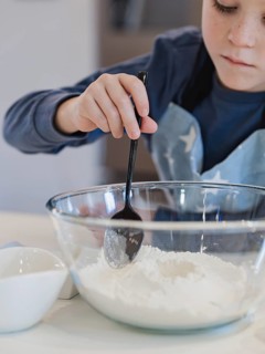 A child mixes flour in a glass bowl using a black spoon in a home kitchen. The scene captures a