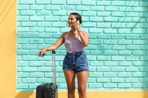 A young Latin woman joyfully stands by a turquoise wall holding her suitcase and chatting on her