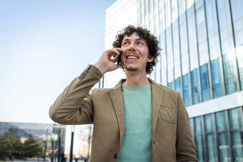 A businessman in a beige jacket enjoys a phone call outside modern glass buildings. He radiates