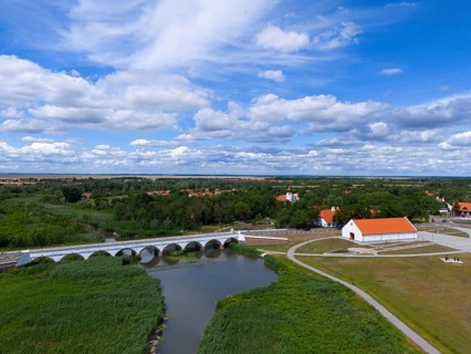 Panoramic view of a river with a bridge and a surrounding village under a wide, cloudy sky, aerial
