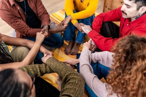 Multiracial group of people are sitting in a circle, holding hands together in a gesture of