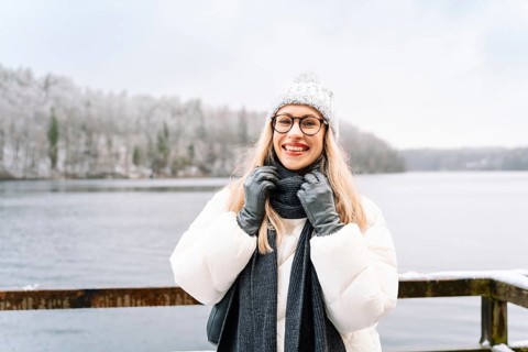 Smiling woman adjusting her scarf on a winter day near a snowy lake. A woman wearing a white winter