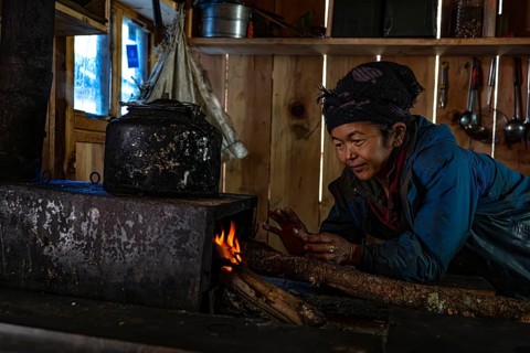 Elderly woman warms herself at a simple cooking place, fireplace with her hands, locals of the