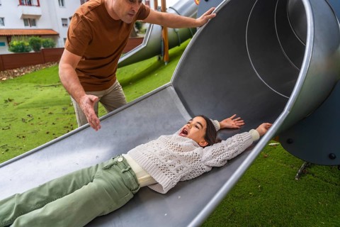 Expressive young girl with excited face sliding down a metal slide while father watches closely,